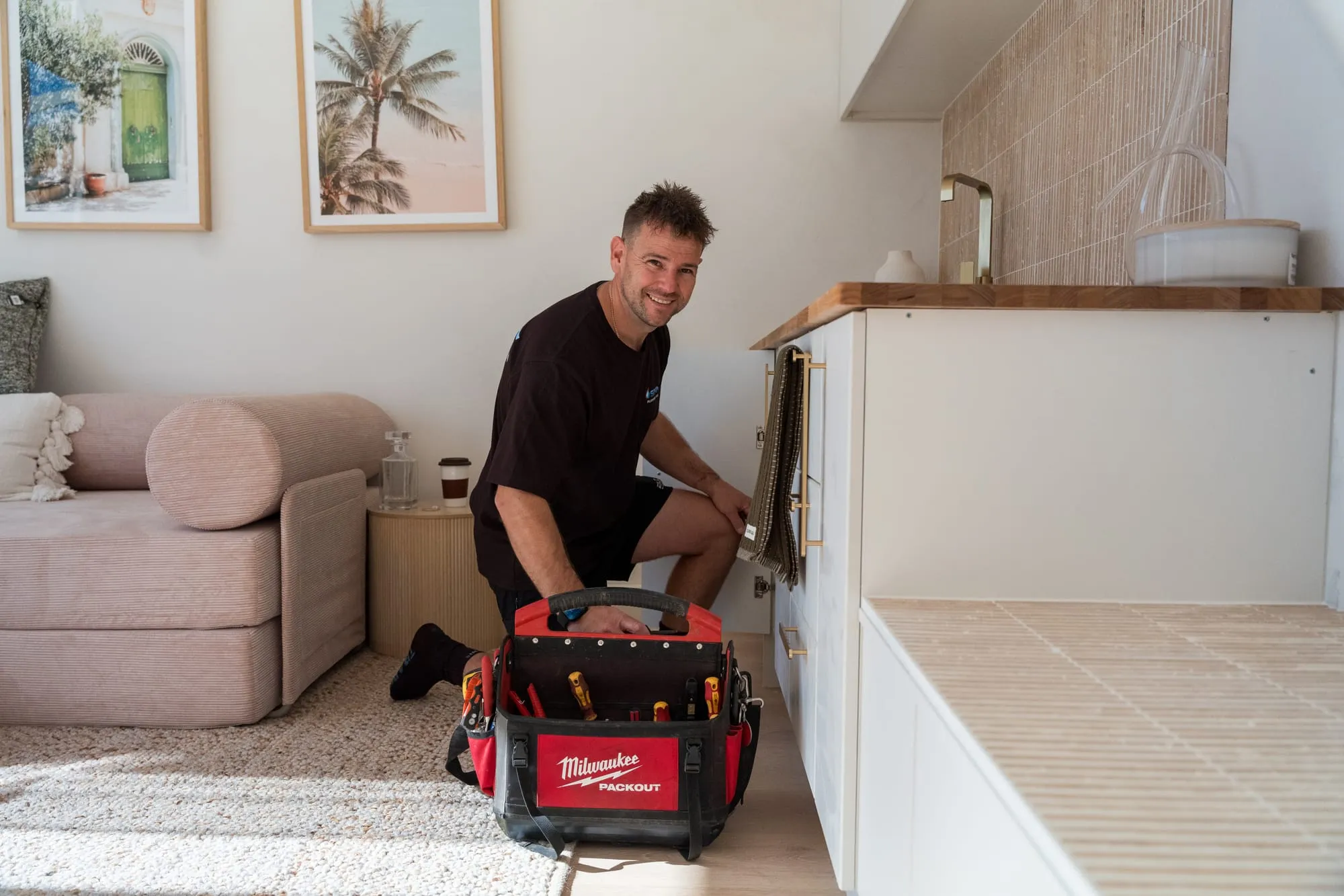 Simms plumber kneeling at a kitchen cabinet with his toolbag open